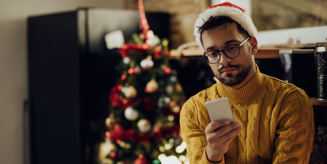 Man in yellow sweater, Santa hat, and Christmas tree in the background holding a phone with a sad expression 