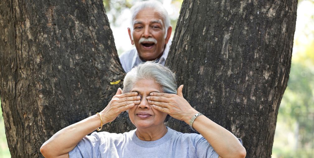 older couple with husband sneaking up on his wife by a tree